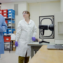 Ryan Falkenstein-Smith and Amy Mensch, both wearing lab coats and gloves, stand in the lab next to a black camera-like device on a table. 