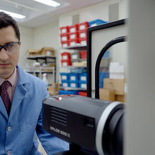 Ryan Falkenstein-Smith, wearing a lab coat and safety glasses, adjusts a black camera-like device in the lab. 