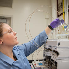 Alix Rodowa wears safety glasses and gloves in the lab as she reaches for a container of clear liquid on top of a piece of scientific equipment. 