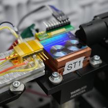 Chip that converts lights into microwave signals. The chip is a fluorescent panel that looks like two tiny vinyl records. A gold box sits to the left of the chip, which is the semiconductor laser.