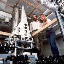 Two smiling researchers stand next to a tall, cylindrical scientific device in a lab.