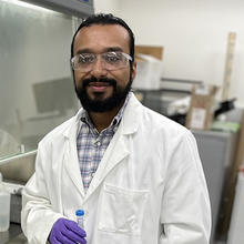 George Caceres wears safety glasses and a lab coat as he poses for a head shot in the lab. 