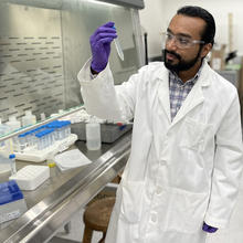 George Caceres wears safety glasses and a lab coat as he looks at a plastic test tube he is holding.