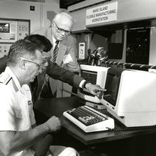 A black and white photograph of a naval officer seated at a computer workstation, another man pointing to the workstation, and a third man standing nearby.