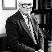 Vertical, black and white image of a man in a suit sitting at the edge of a desk.