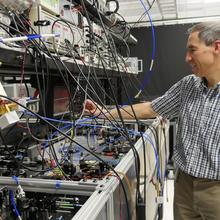 Tasshi Dennis stands in his lab and reaches toward a countertop full of equipment with lots of wires and circuitry.