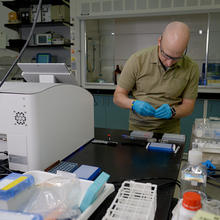Thomas Forbes stands at a lab table near a white plastic 3D printer, looking down at materials he is preparing. 