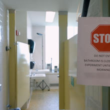 A paper sign is clipped in front of a open door to a public restroom. The sign reads, “Stop. Do Not Enter. Bathroom is closed for an experiment until Monday Morning.”