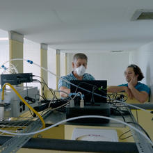 A jumble of wires, tubes, and electronics rest on impromptu shelves placed across the walls of a bathroom stall. In the background, two men look at a laptop together.