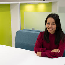 Mei Lee Ngan poses smiling and sitting at a white table with a bright green wall behind her. 