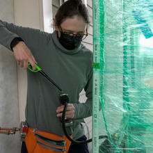 A researcher standing outside a house holds a lighter to the end of a hose-like device leading into an area closed off by plastic wrap. 