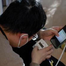 A researcher bends over a digital readout on a piece of equipment, comparing it to information on his phone. 