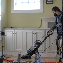 A researcher runs a vacuum cleaner over the wood floor in a living room that's empty except for scientific equipment and wiring. 