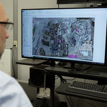 View from behind shows two seated researchers looking at a computer screen that shows an aerial view of a large outdoor site. 
