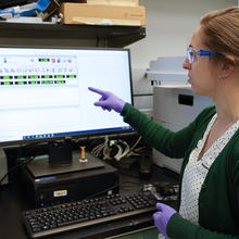 Briana Capistran wears safety glasses and purple gloves as she points to a computer screen showing information from lab analyses. 