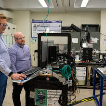 Two researchers in safety glasses stand looking at a computer screen in a lab filled with equipment.