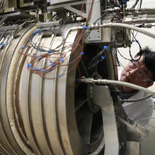 A researcher peers around the edge of large cylindrical piece of equipment in the lab. 