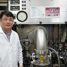 A researcher in a lab coat and safety glasses poses next to a large piece of lab equipment.