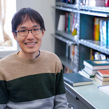 Leroy Jia poses smiling at his desk, with piles of books in the background. 