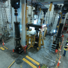 A high-ceilinged lab space is filled with large pieces of equipment, including a square structure in the center. A few people in hard hats stand at the edge of the room. 