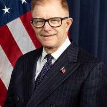 Headshot of a man wearing glasses and a dark suit with an American flag behind him