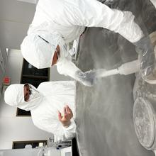 Researchers in white hazmat coveralls reach into an open freezer with a scooping device as vapor rises from the freezer.