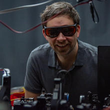 Krister Shalm, wearing safety glasses, smiles over a lab table covered with electronics equipment.