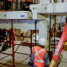 A person wearing construction safety gear looks up at a cracked concrete slab and beam arrangement held up by scaffolding in a large indoor space. 