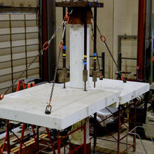 View from above shows cracked concrete slab attached by chains to column and held up from below by scaffolding in a large indoor space. 
