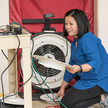 Lisa Ng kneels, holding parts of a circular fan built into a room divider. 