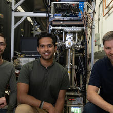 Three male researchers pose smiling in front of a large, complex scientific device in a lab.