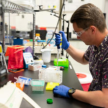 Researcher in safety glasses and gloves sits at a lab bench working with small test tubes.