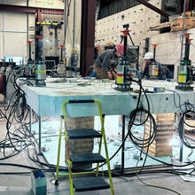 Two researchers in safety gear examine a slab of concrete set up horizontally in a large lab surrounded by scientific equipment.