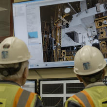 Two people in hard hats, shown from behind, look at a large screen showing concrete slabs and testing equipment.