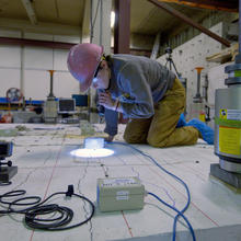 A researcher in a hard hat kneels on a concrete slab and uses a flashlight to examine it closely.