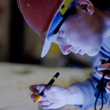 Close-up shows a female researcher in a hard hat looking down, shining a flashlight and holding a marker.