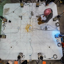 An overhead view shows a researcher in a hard hat making notes while kneeling on a concrete slab surrounded by scientific equipment.