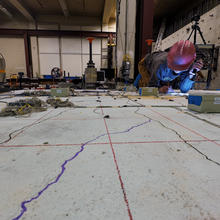 A researcher in a hard hat is making notes while kneeling on a concrete slab surrounded by scientific equipment.