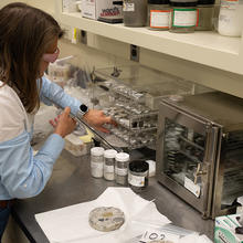A researcher wearing safety glasses leans over the lab table near where a small disk of concrete is sitting. 