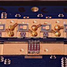 Circuit board with various square chips and gold screws. Small letters in the center say: NIST True Bq.