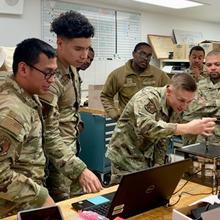 Airmen in uniform gather around a desk that holds an open laptop and several electrical devices, including one inside a clear box. 