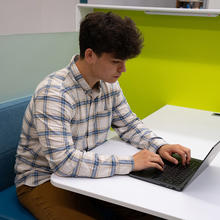 A man works on a laptop sitting at a white table in front of a brightly colored wall. 