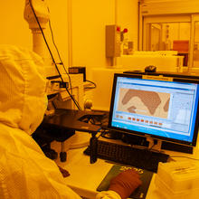 In a lab with orange lighting, a scientist wearing a full suit of protective gear sits at a table with a microscope and looks at images on a screen.
