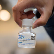 A hand holds a small, clear bottle full of a liquid solution with a label that says SRM 1989.