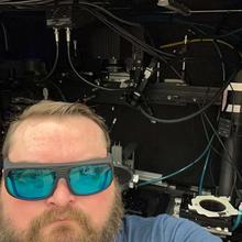 A bearded man wearing reflective safety glasses stands in front of an open cabinet full of scientific equipment. 