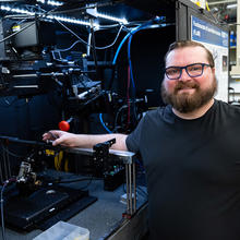 A bearded man poses smiling next to an open cabinet of equipment in a lab. 