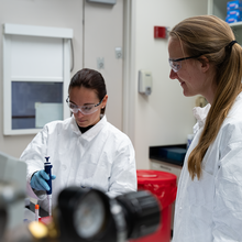 A researcher in a lab coat and safety glasses uses lab equipment as another researcher looks on. 
