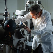 Andrew Iams wears a lab coat and safety glasses as he looks into a circular opening on a large piece of lab equipment. 