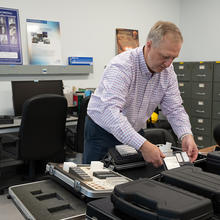 In an office with a desk and filing cabinets, Jack Glover leans over a table holding black cases with customized foam inserts for equipment. 