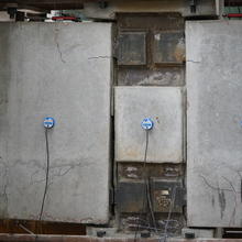 A wide view a concrete beam assembly inside a large metal frame. There are multiple small cracks in the concrete highlighted with Sharpie. One of the steel plates connecting the concrete pieces together is broken.